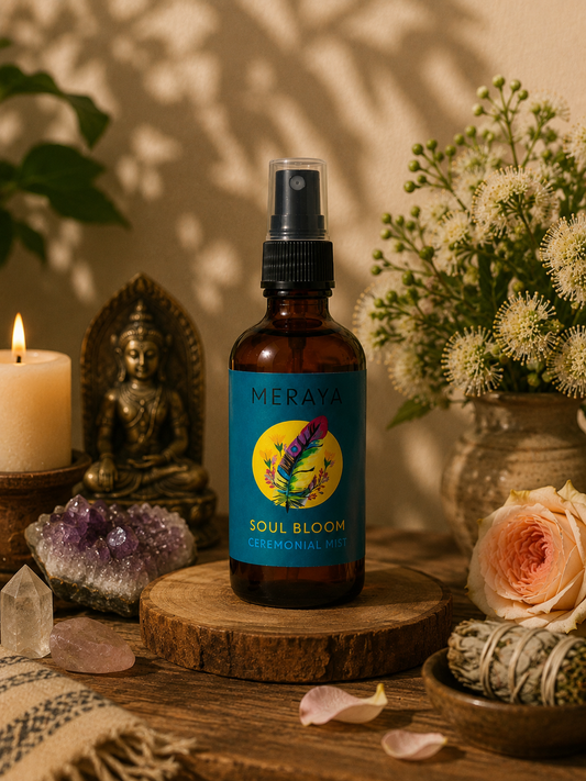 A bottle of Meraya Soul Bloom Emotional Support Spray stands on a wooden slice in a styled setting with a lit candle, bronze Buddha statue, crystal cluster, flowers, and a rose.