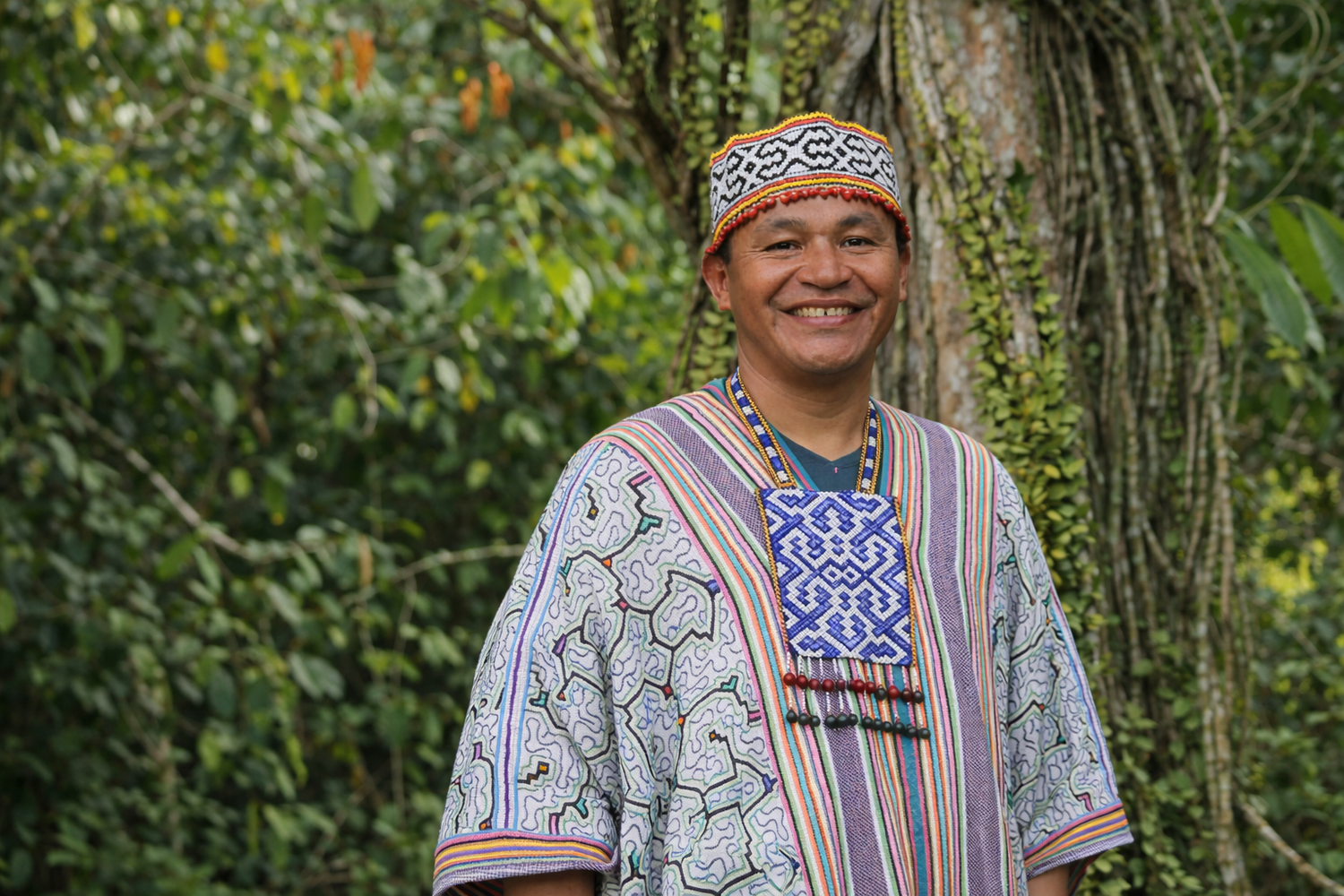 Maestro wearing traditional shipibo attire with a colorful pattern, standing in front of green foliage.