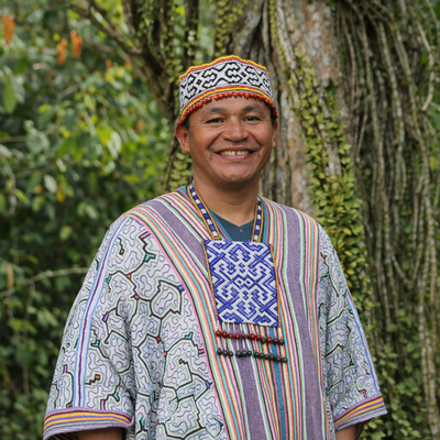 Maestro wearing traditional shipibo attire with a colorful pattern, standing in front of green foliage.