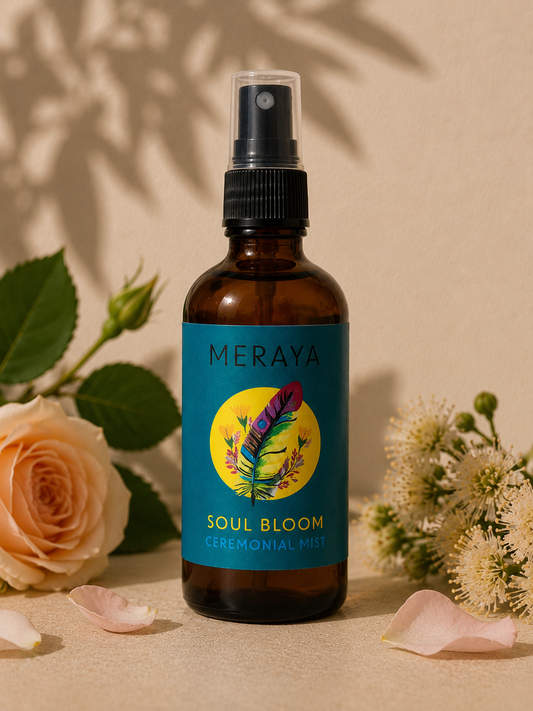 Meraya Soul Bloom Ceremonial Mist in an amber glass spray bottle with a teal label, styled with peach roses and white flowers on a beige surface with warm sunlight and leaf shadows.